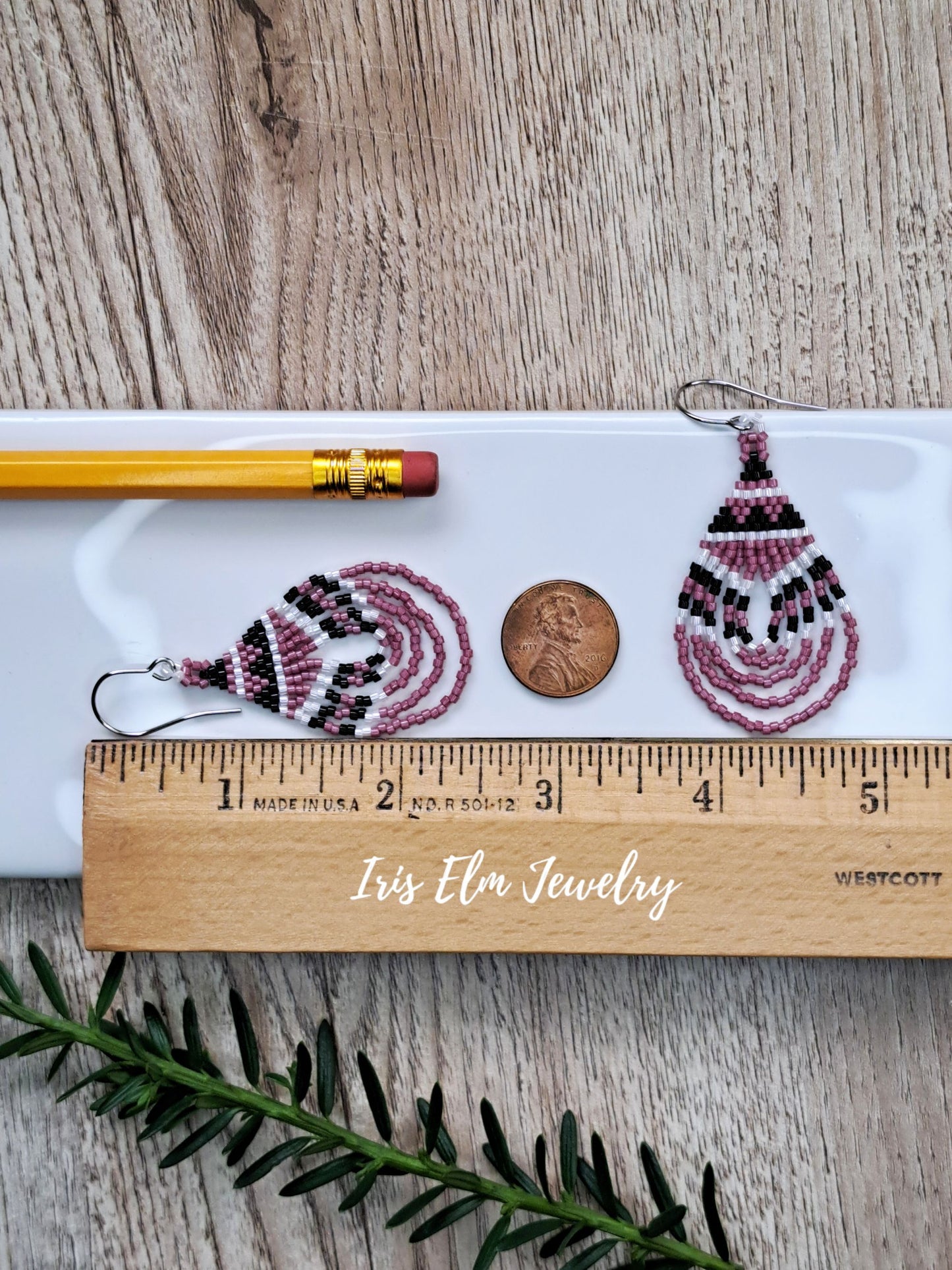 A pair of 2.5 inch long handmade beaded teardrop earrings by Iris Elm Jewelry, shown on a white tray with a pencil, a penny, and a wooden ruler on a wood-grain background.