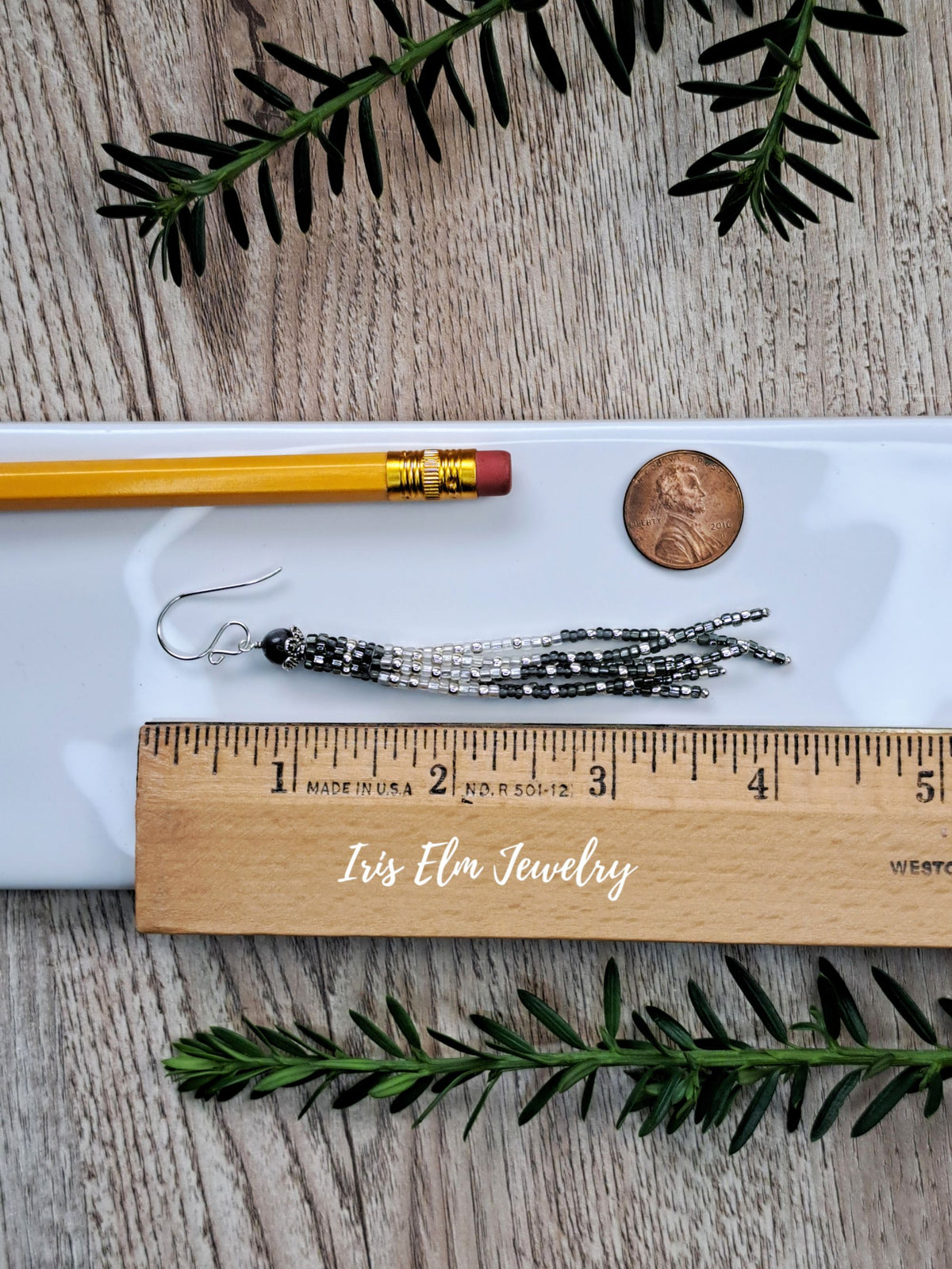 A pair of 4 inch long handmade beaded tassel earrings in silver and black lies on a white tray, styled with a wooden ruler, pencil, penny, and pine branches on a rustic wood background. The brand name 'Iris Elm Jewelry' is printed on the wooden ruler.