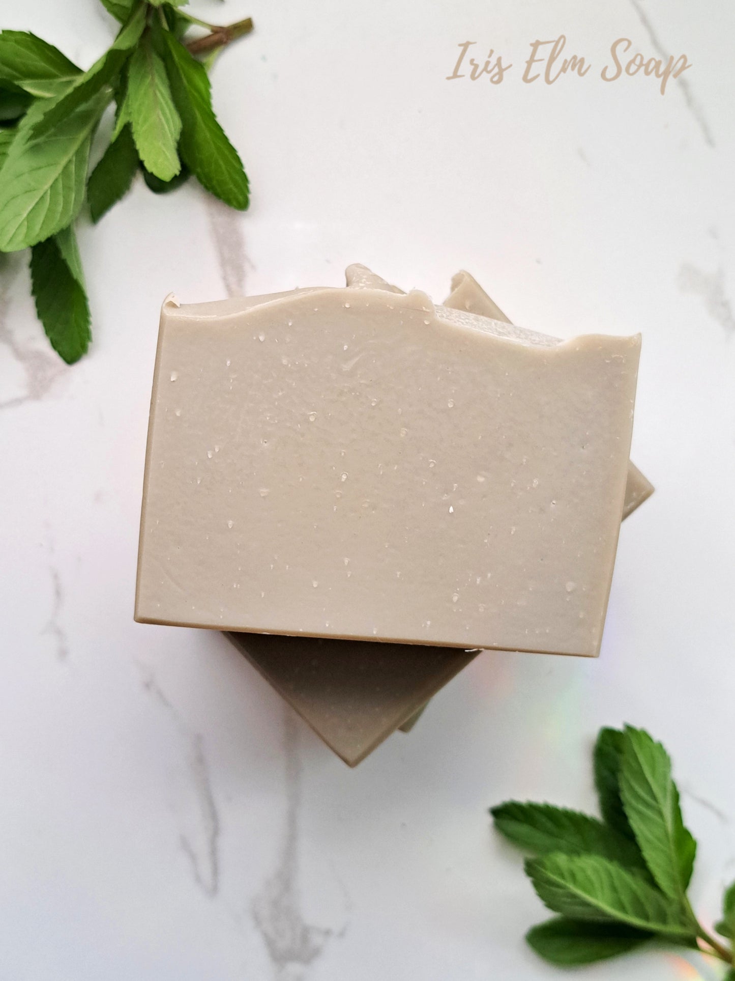 Two eucalyptus tea tree mint handmade essential oil soap bars on a white marble surface with green leaves around, featuring 'Iris Elm Soap' branding.