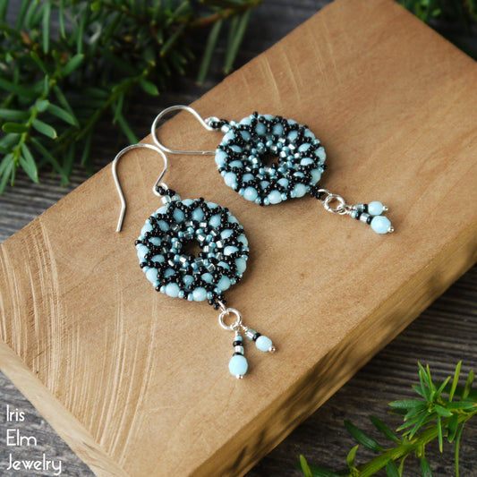 Beaded light blue Czech glass wreath earrings with black accent beads on a wooden block with greenery in the background