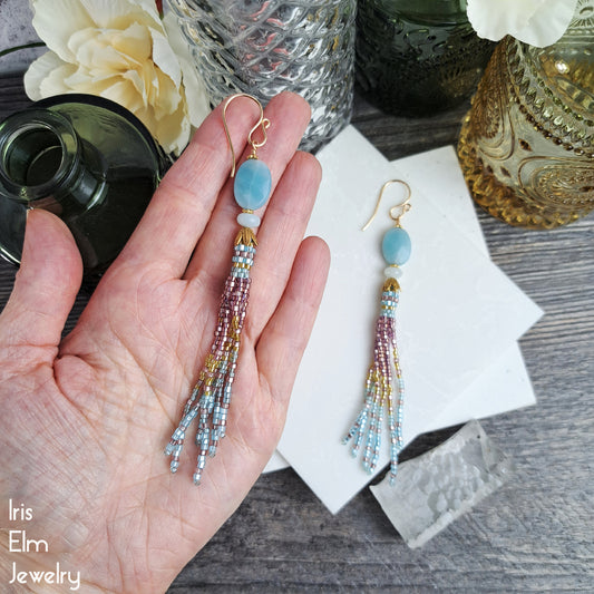 Beaded pink, gold, and blue amazonite stone and seed bead fringe tassel earrings held in a hand with a decorative background