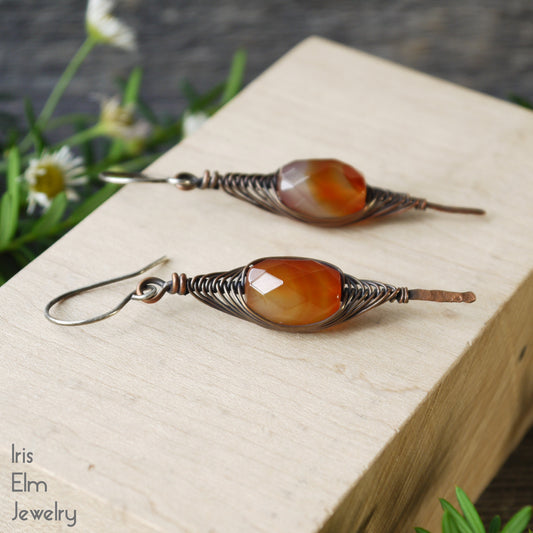 Pair of orange red agate gemstone copper wire wrapped herringbone earrings on a wooden block with 'Iris Elm Jewelry' branding.