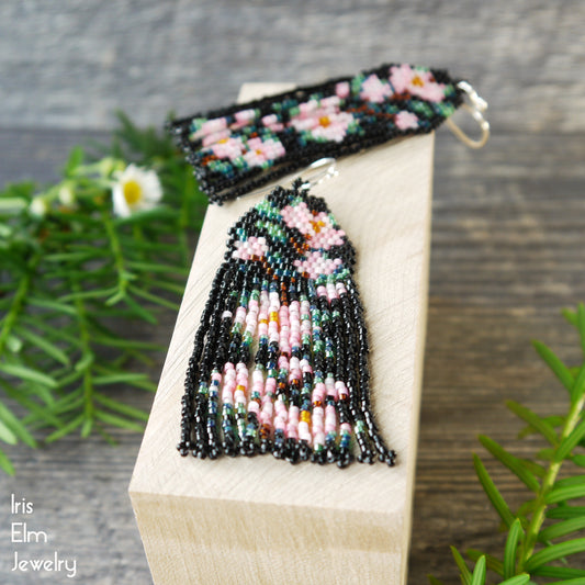 Beaded pink cherry blossom flower fringe seed bead earrings on a wooden block with plants in the background, featuring the brand 'Iris Elm Jewelry'.
