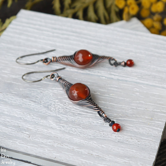 Pair of oxidized copper wire wrapped herringbone earrings with red agate stones on a textured white surface