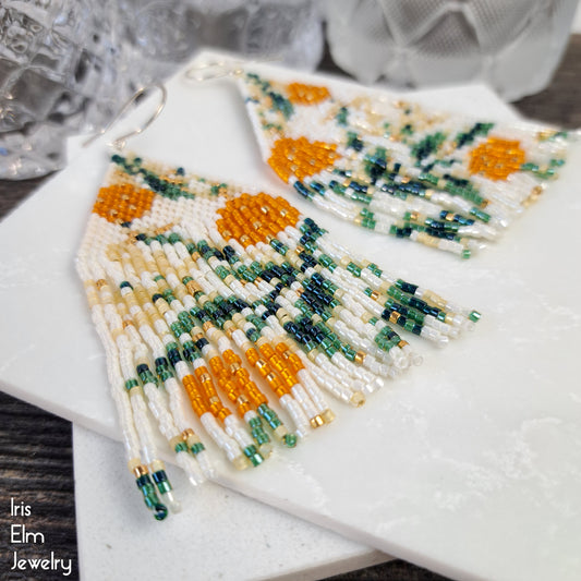 Beaded earrings with orange and green floral pattern on a marble surface, brand 'Iris Elm Jewelry' visible.