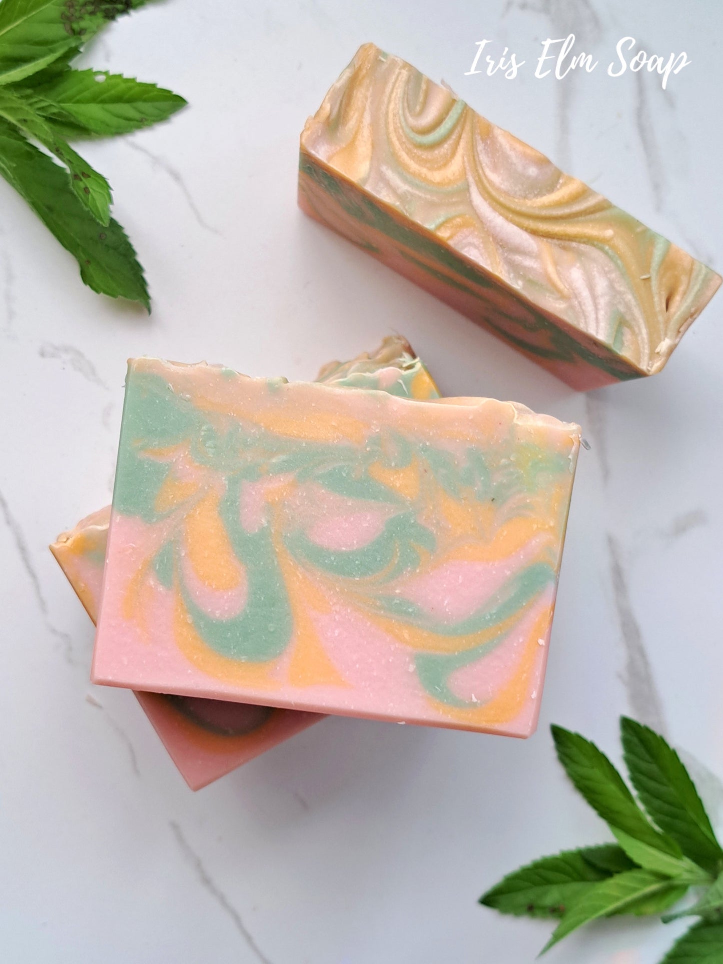 Two swirled grapefruit and mint handmade soap bars on a white surface with green leaves.
