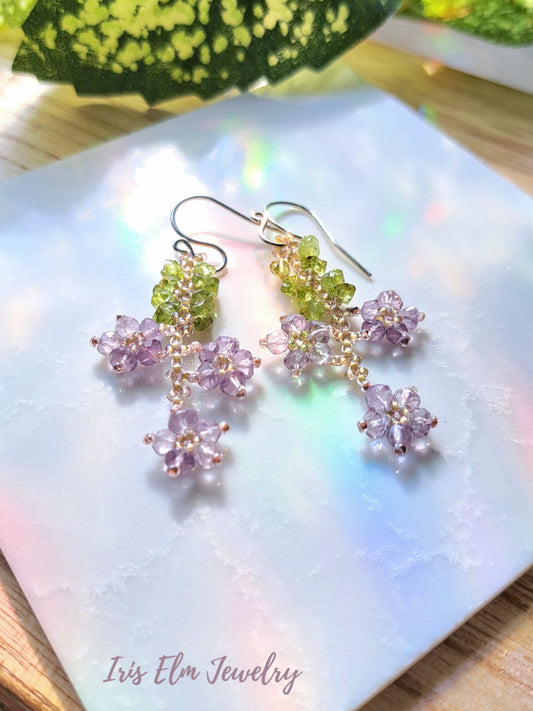 Pair of floral earrings with purple amethyst and green peridot stones on a light fabric background, branded 'Iris Elm Jewelry'.