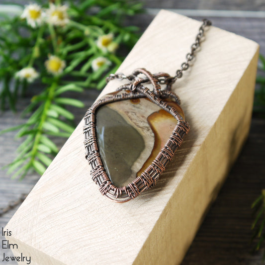 Necklace with a jasper handmade stone wire woven copper pendant on a wooden block, with plants in the background