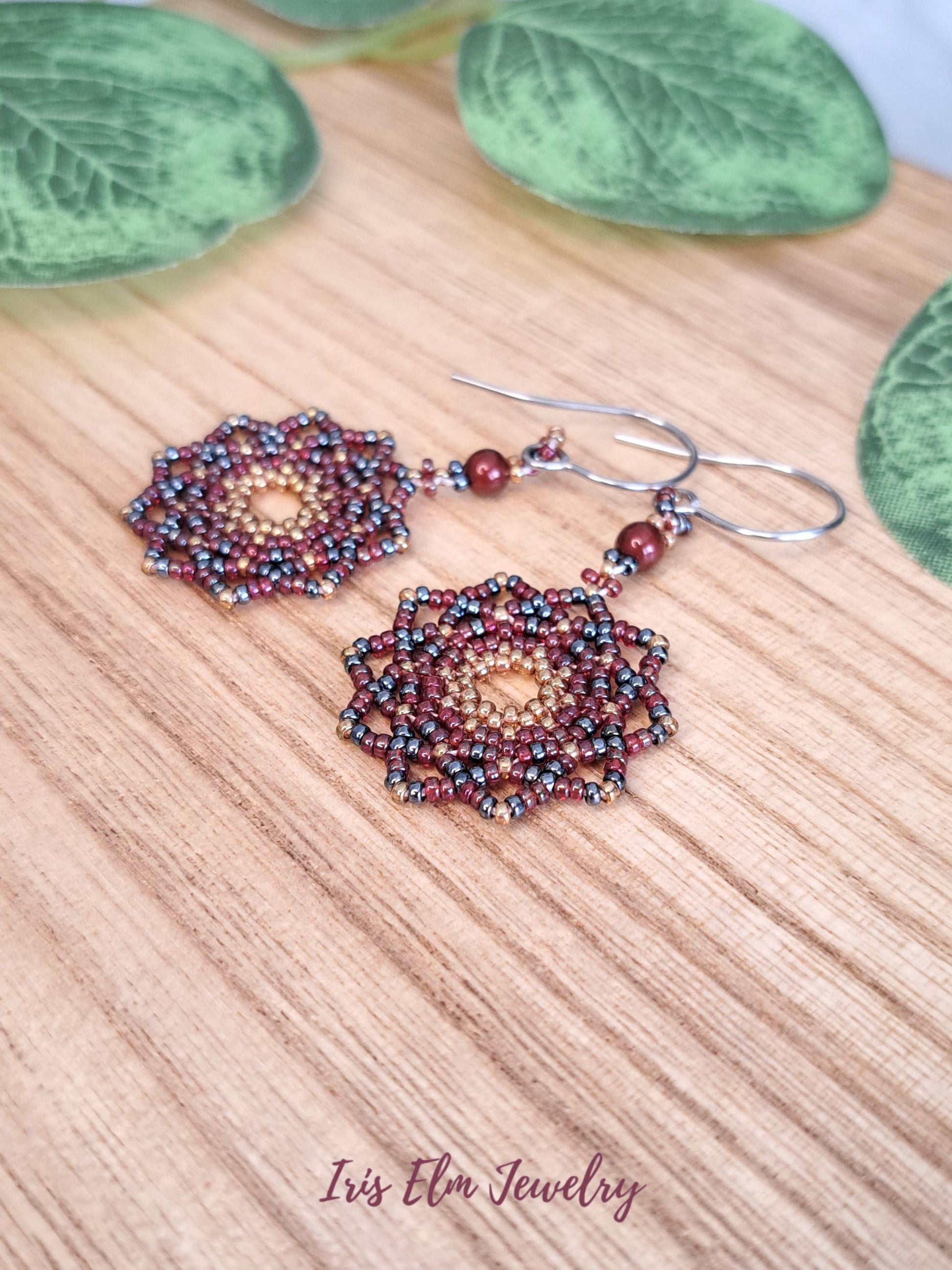 Beaded red woven glass bead mandala earrings on a wooden surface with green leaves in the background, featuring 'Iris Elm Jewelry'.
