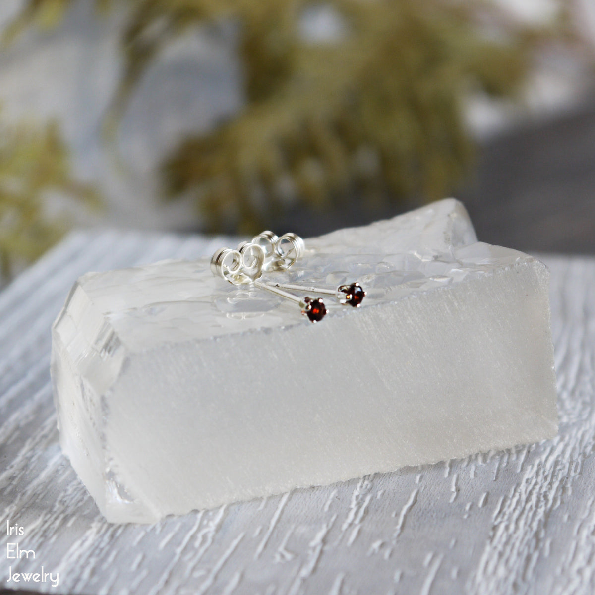 Pair of silver 2mm stud earrings with red garnet gemstones on a white stone surface, brand 'Iris Elm jewelry' visible.