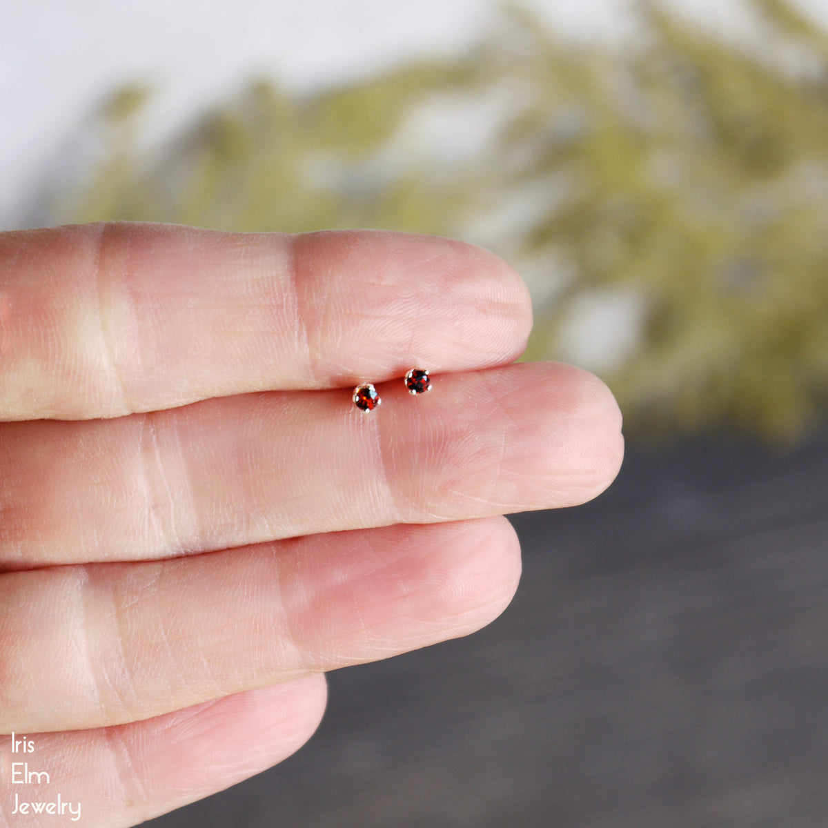 Small garnet gemstone red earrings held between two fingers with a blurred natural background