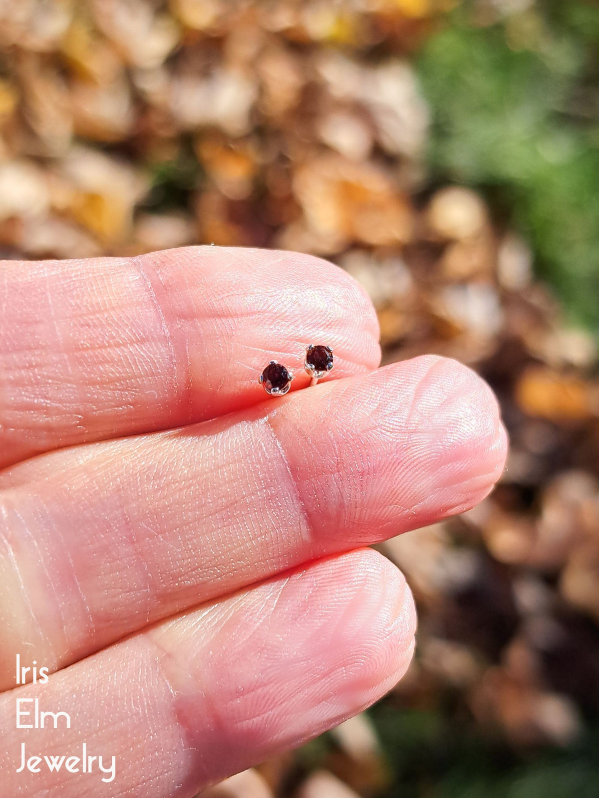 tiny 2mm sterling silver garnet stud earrings held between fingers with blurred natural background