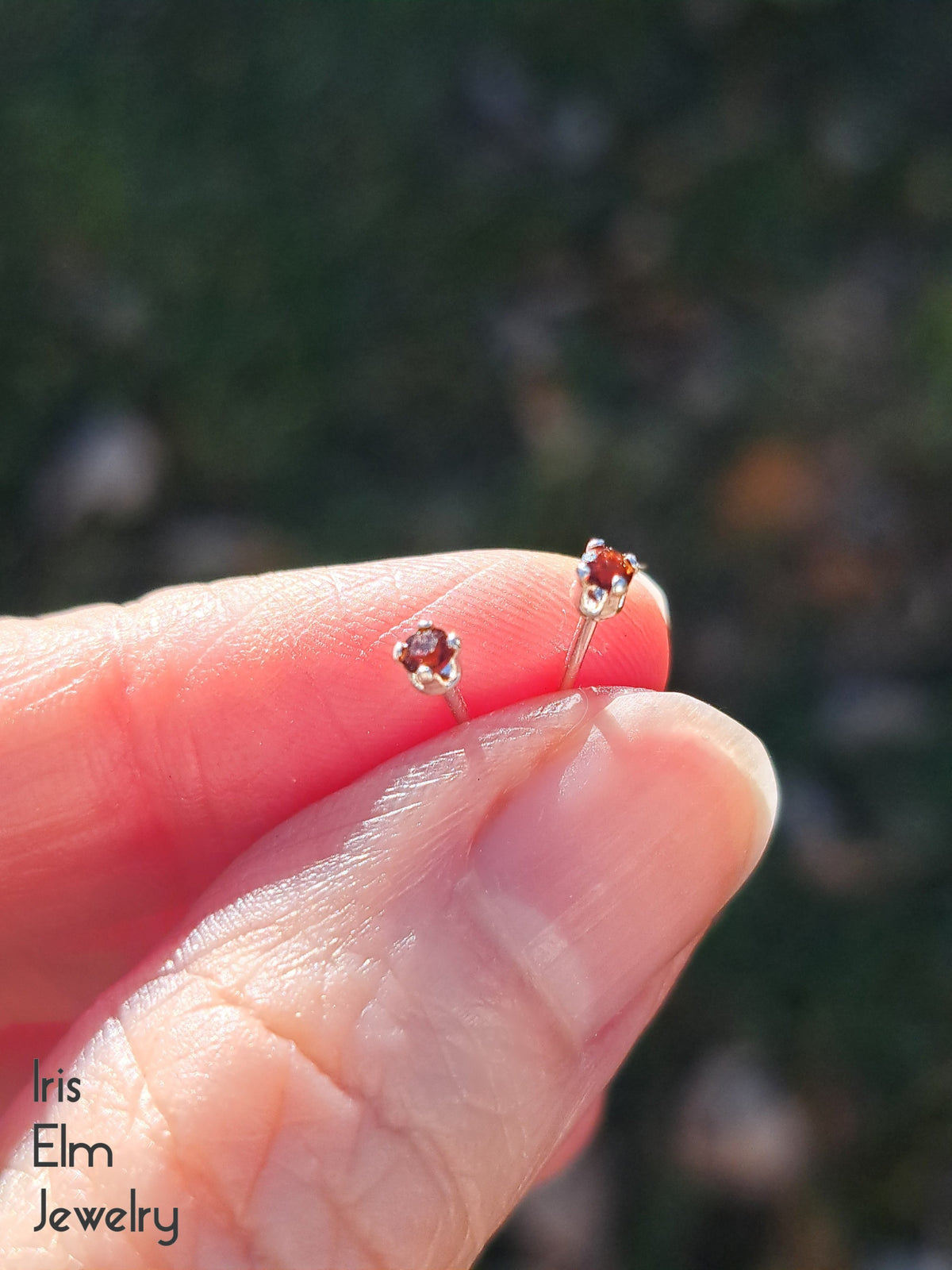 Two small garnet post earrings being held between fingers with a blurred natural background