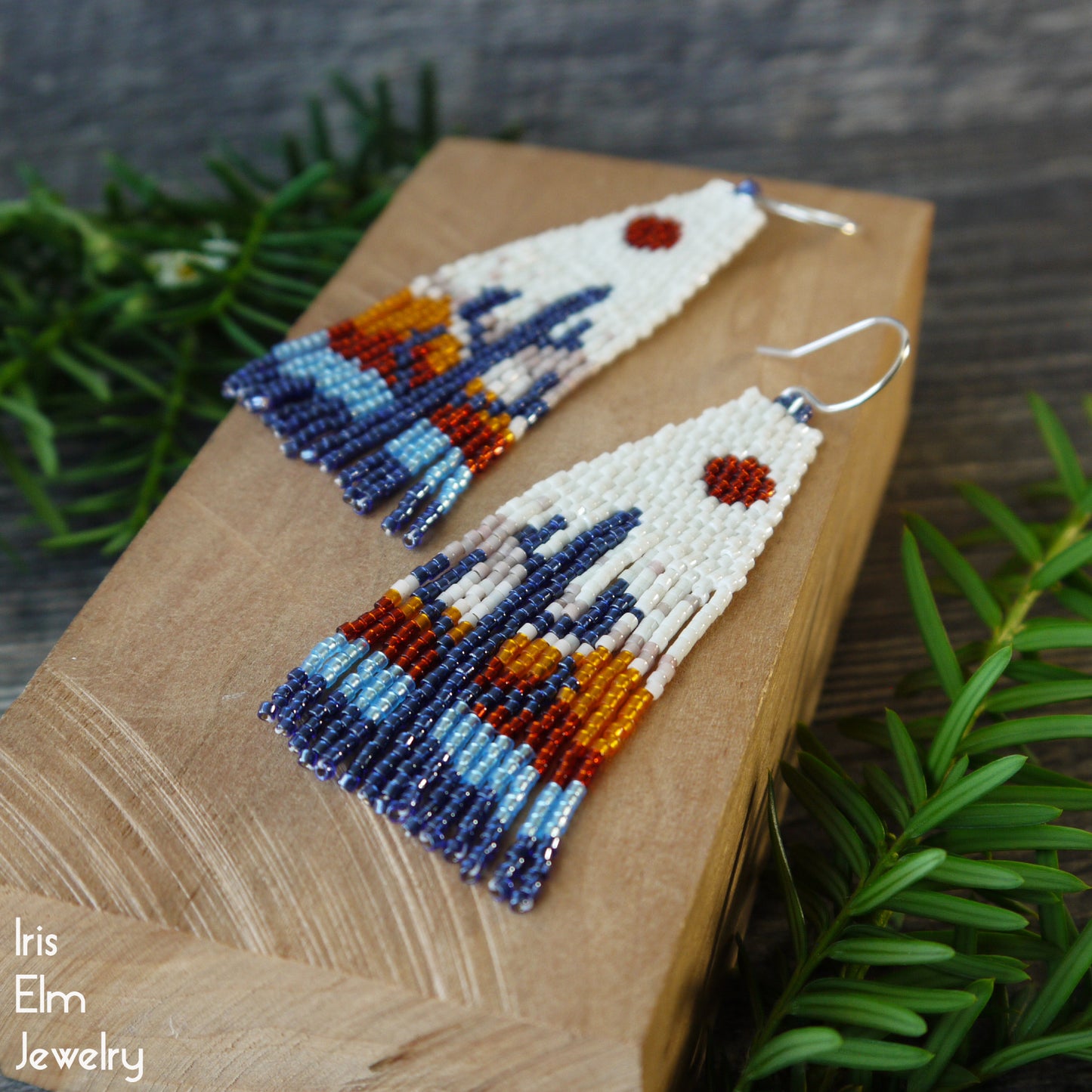 Beaded seed bead fringe earrings with sunset cactus design on a wooden surface with greenery in the background, featuring the brand 'Iris Elm Jewelry'.