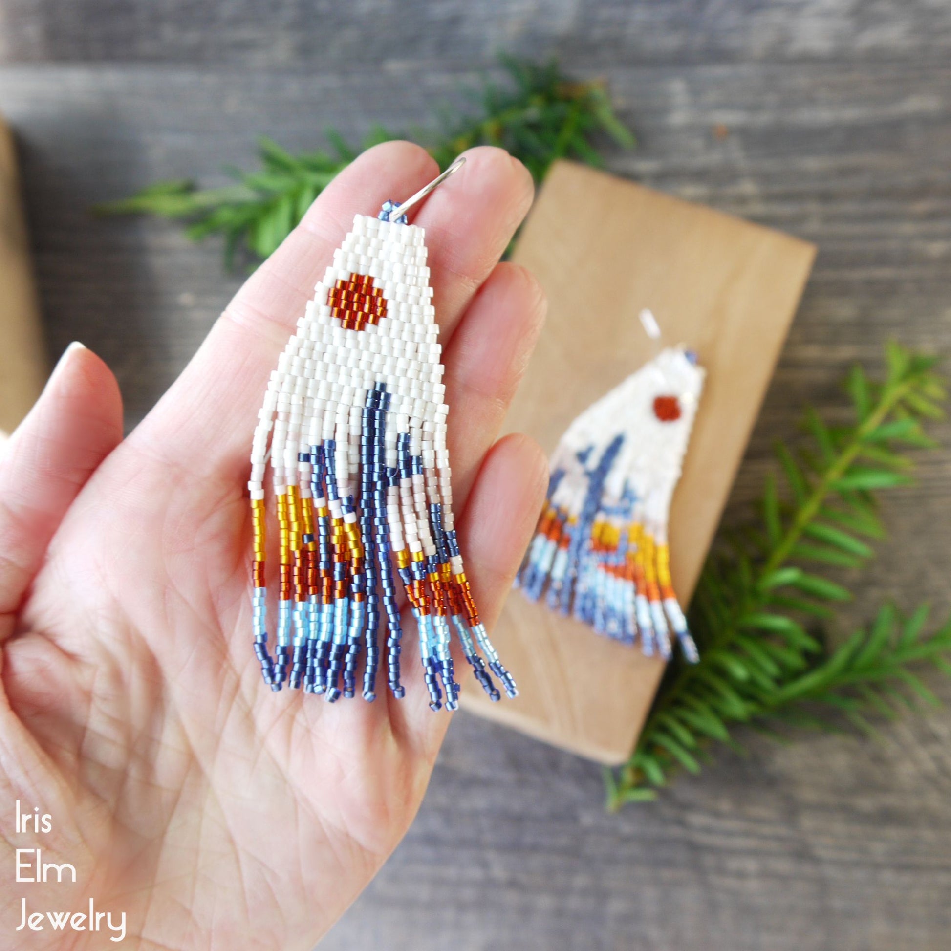 Hand holding beaded cactus fringe blue and orange earrings with a wooden block and greenery in the background, featuring 'Iris Elm Jewelry'.