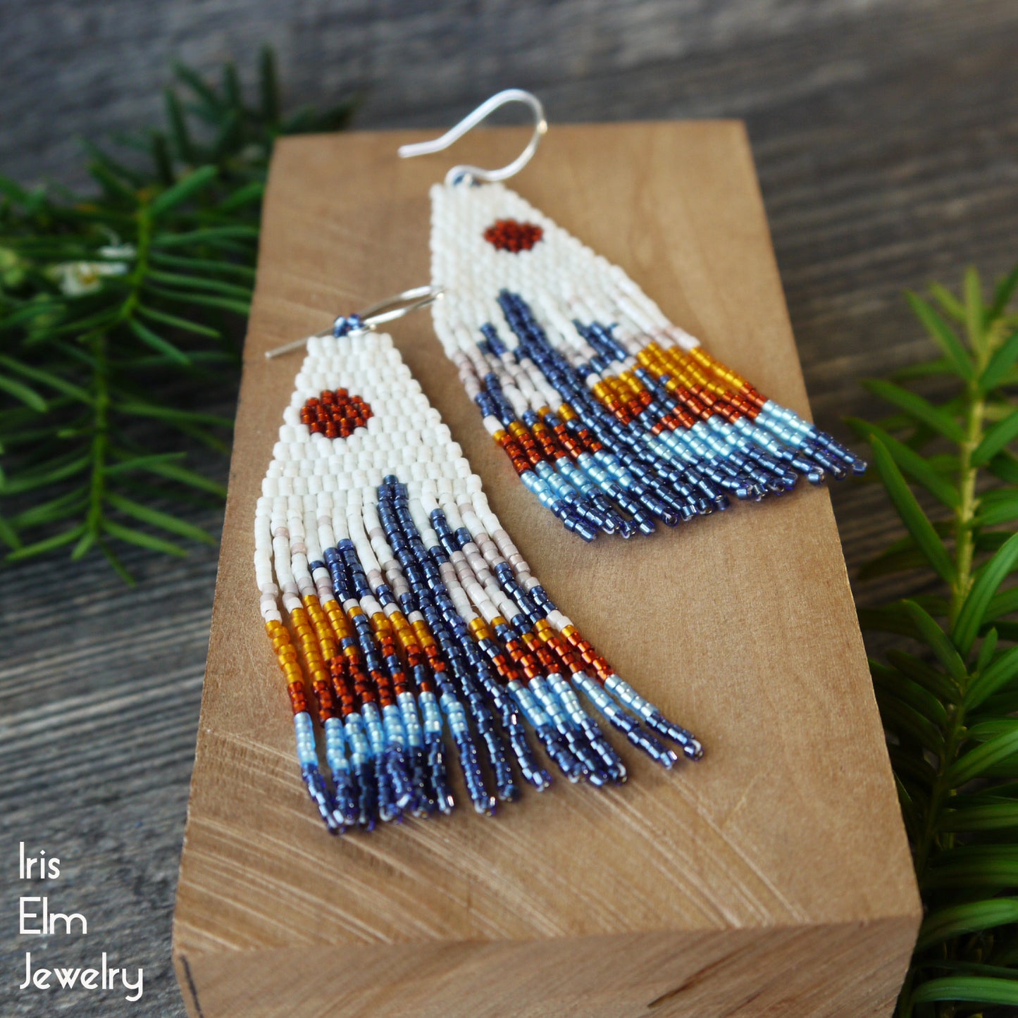 Pair of handmade beadwork seed bead cactus fringe sunset earrings on a wooden block with greenery in the background, featuring the brand 'Iris Elm Jewelry'.