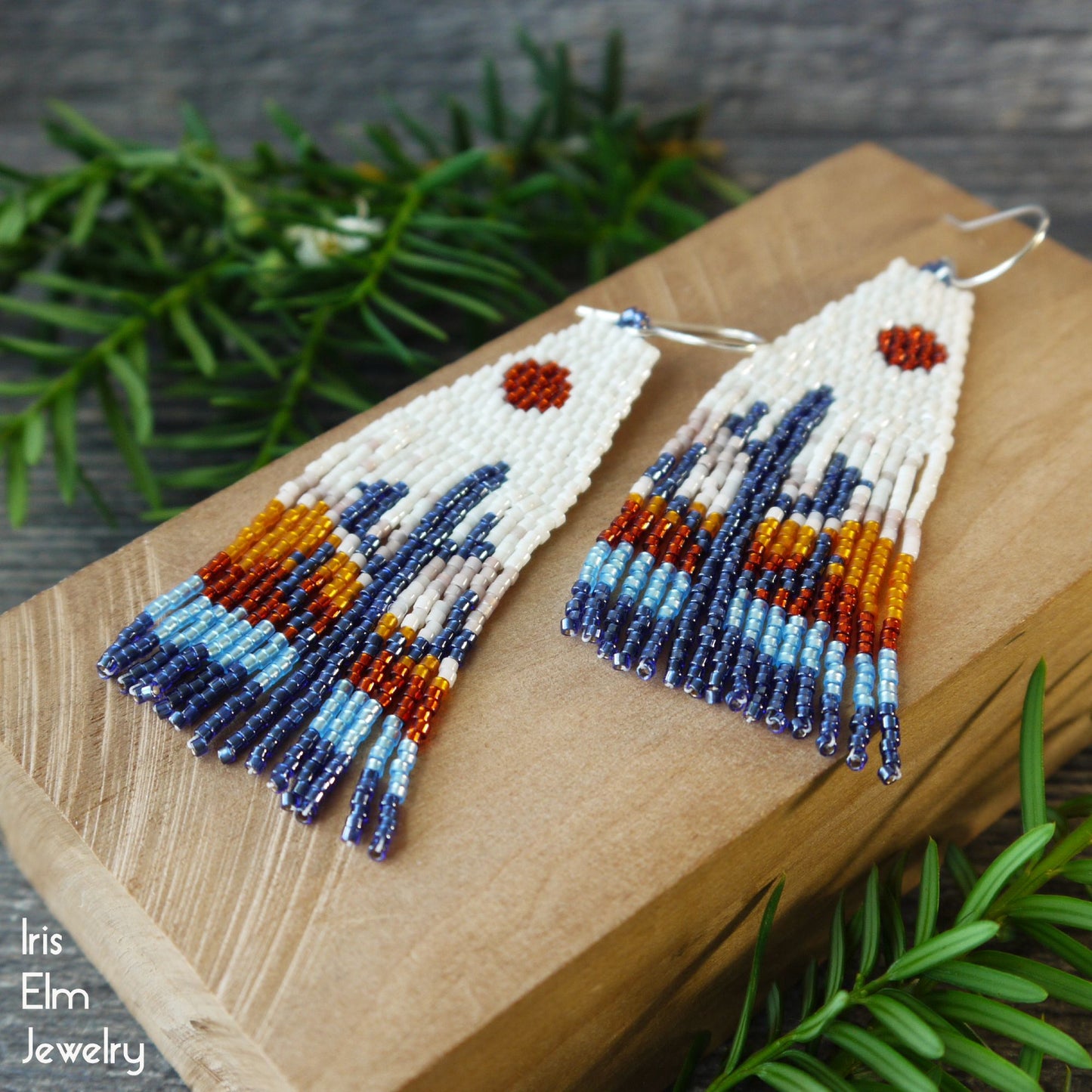 Beaded fringe earrings with sunset cactus design on a wooden block with green foliage in the background