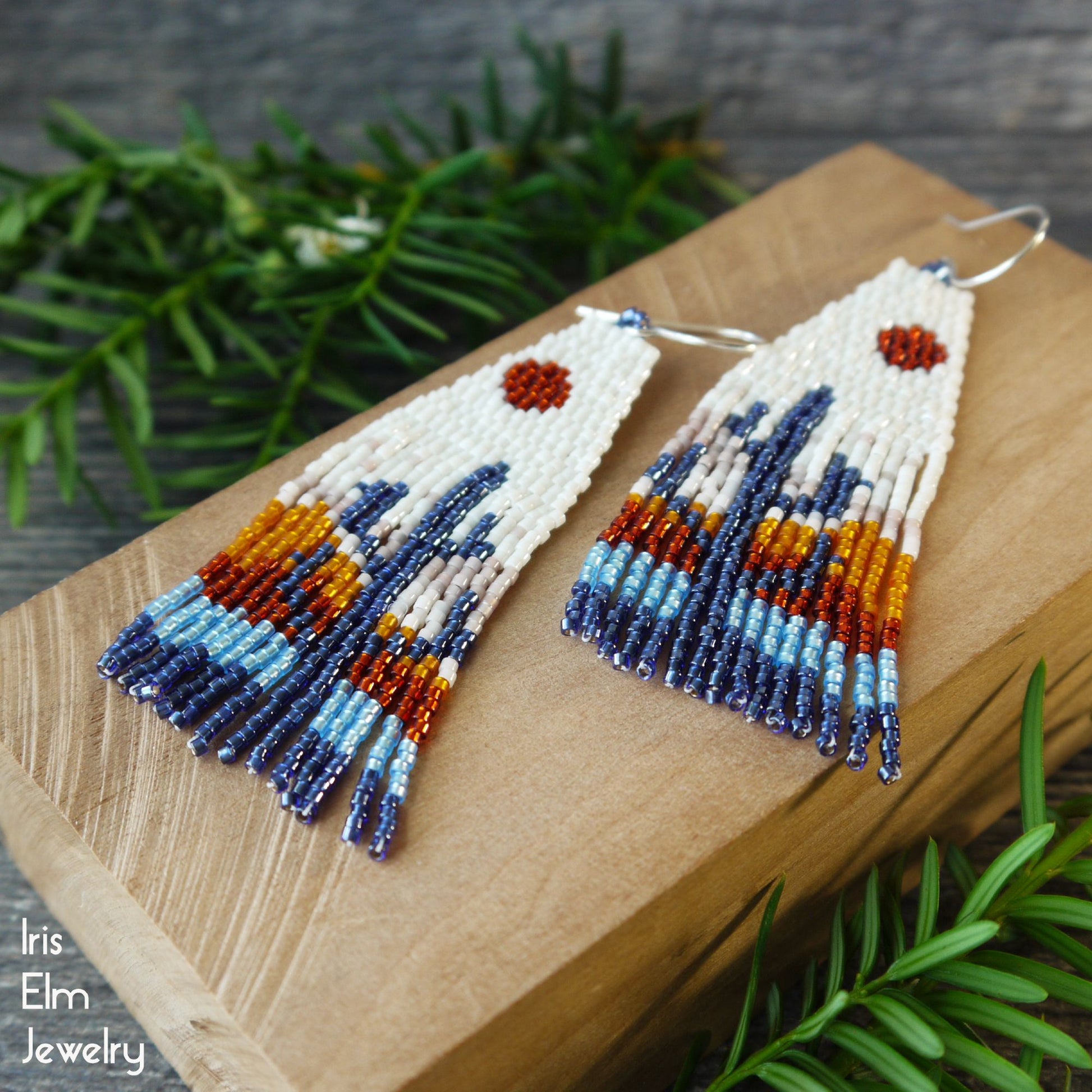 Beaded fringe earrings with sunset cactus design on a wooden block with green foliage in the background