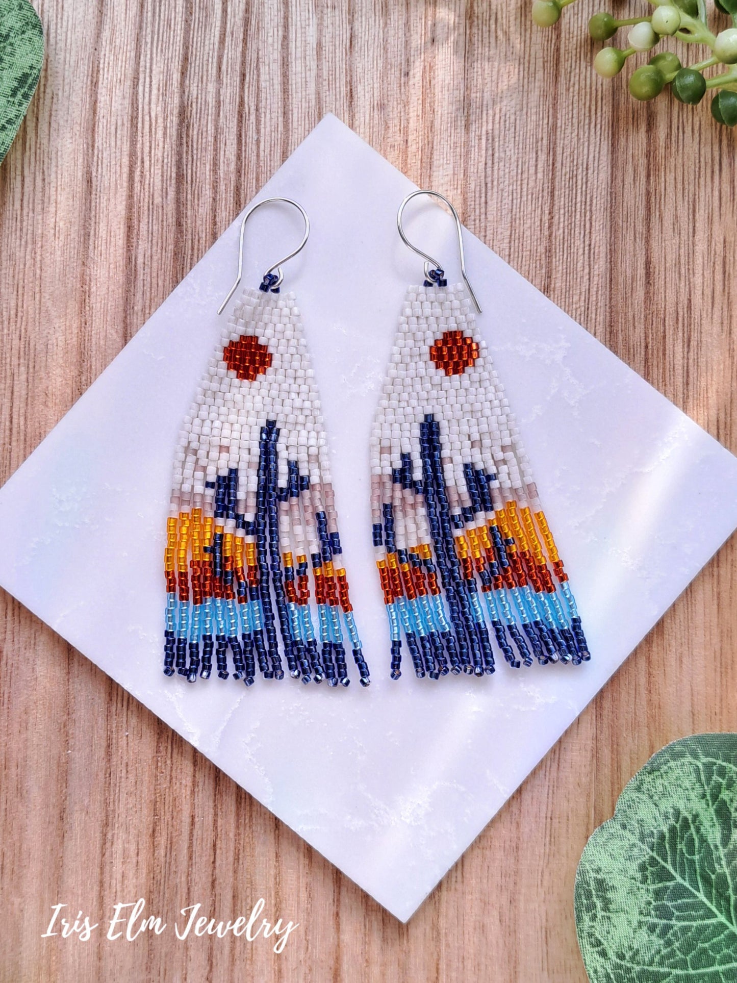Beaded desert sunset cactus seed bead fringe earrings on a white square with 'Iris Elm Jewelry' branding, placed on a wooden surface.