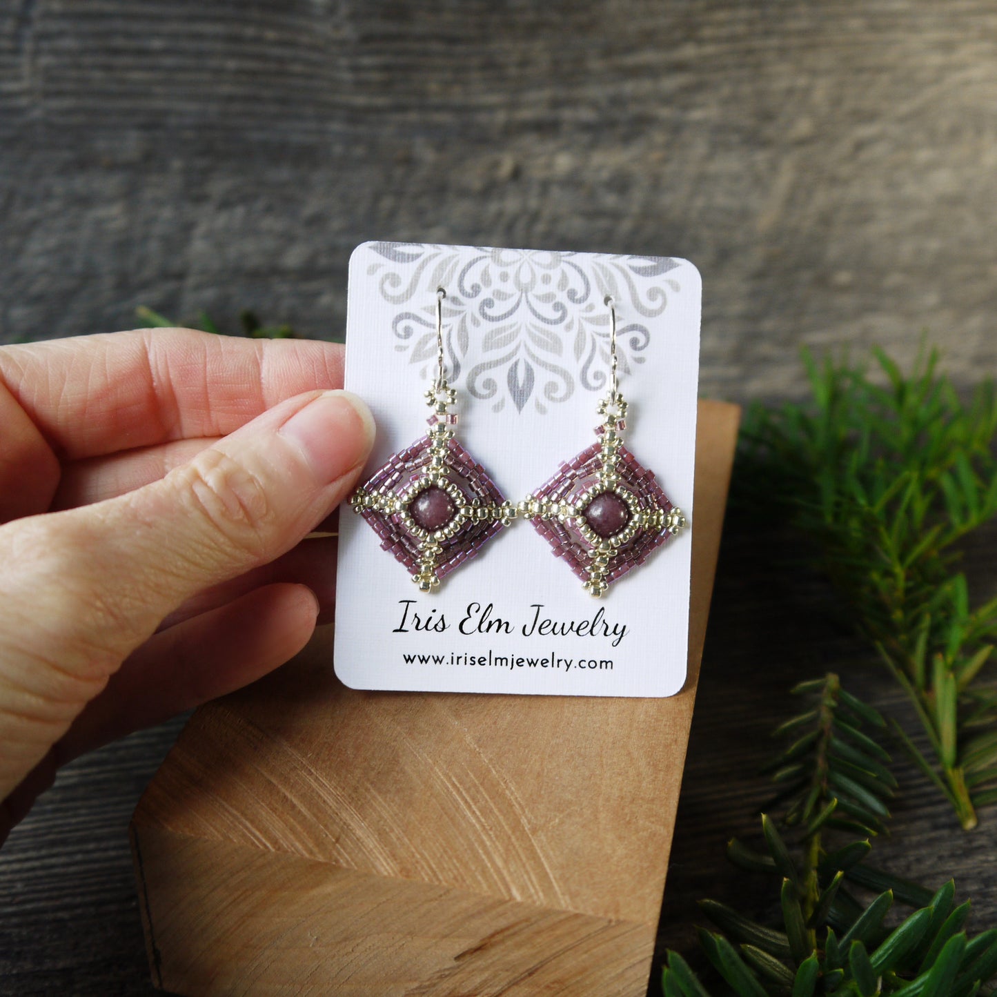 Hand holding a pair of beadwoven lepidolite earrings on an Iris Elm Jewelry card with a wooden surface and plant in the background.