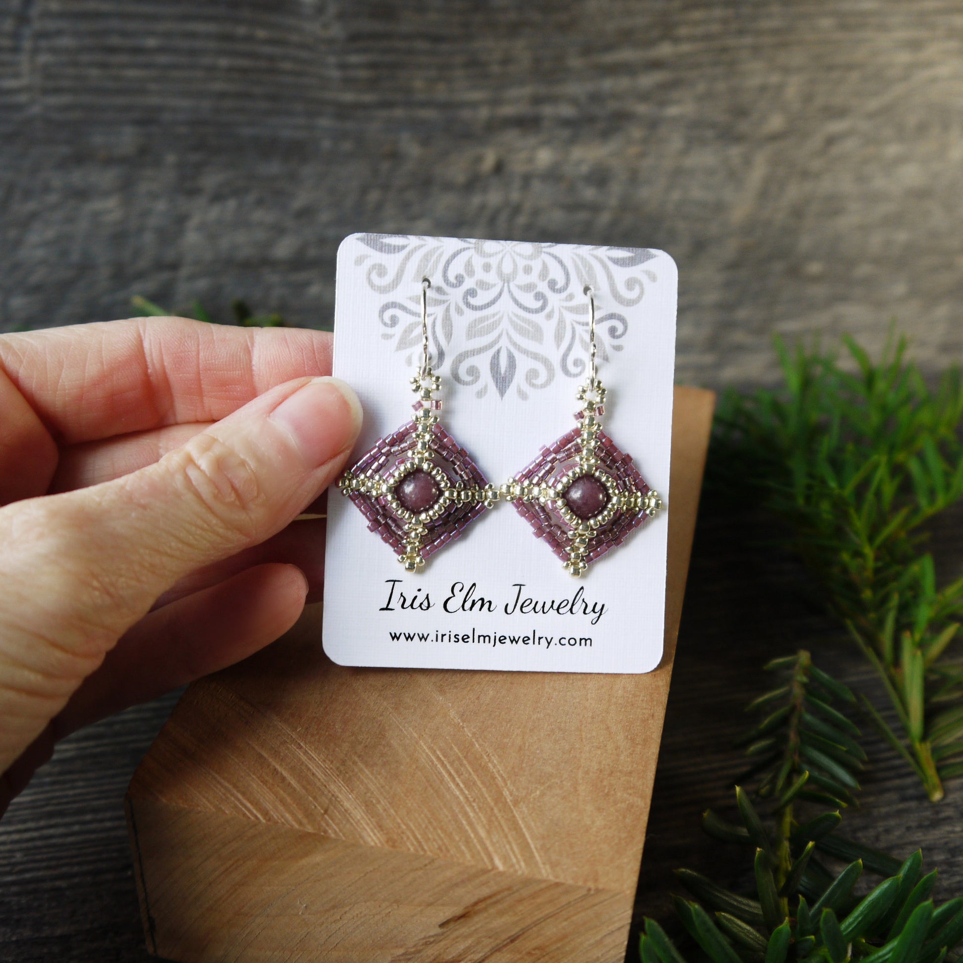 Hand holding a pair of beadwoven lepidolite earrings on an Iris Elm Jewelry card with a wooden surface and plant in the background.