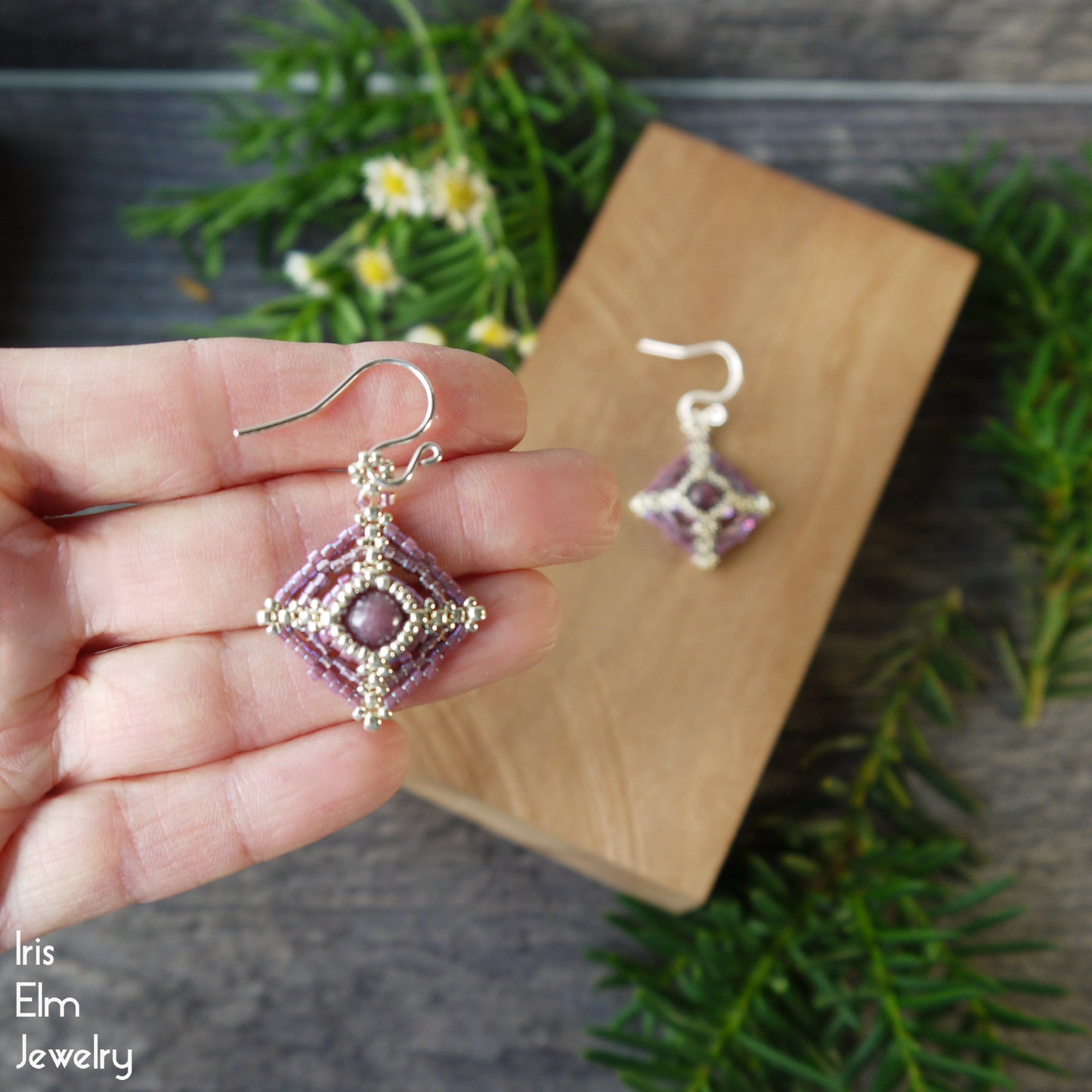Hand holding a pair of beaded earrings with lepidolite stone surround by seed bead beadwork with a wooden display board in the background.