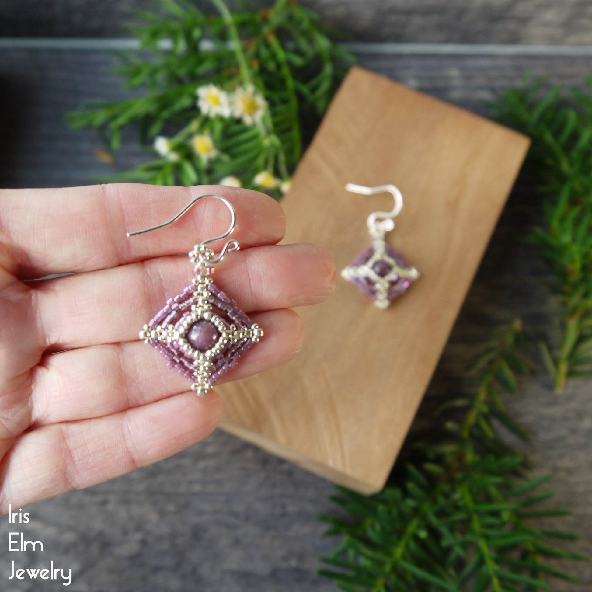Hand holding a pair of beaded earrings with lepidolite stone surround by seed bead beadwork with a wooden display board in the background.