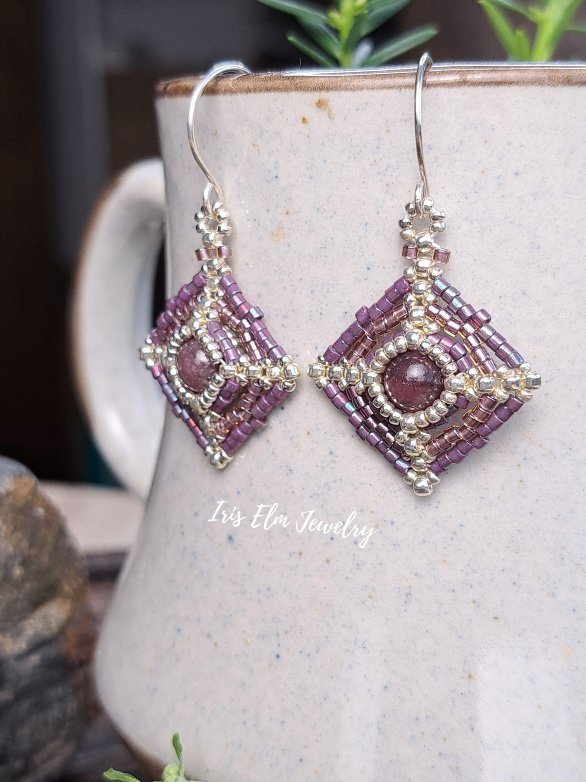 A pair of handmade beaded dangle earrings with purple and silver details, displayed on a speckled ceramic mug with a watermark reading 'Iris Elm Jewelry'.