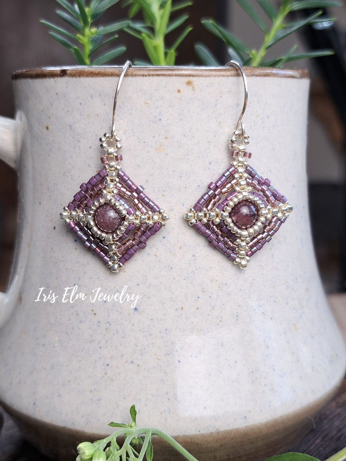 A pair of handmade beaded dangle earrings with purple and gold geometric patterns, hanging from silver fishhook wires. The earrings are displayed against a speckled beige ceramic mug with green foliage in the background.