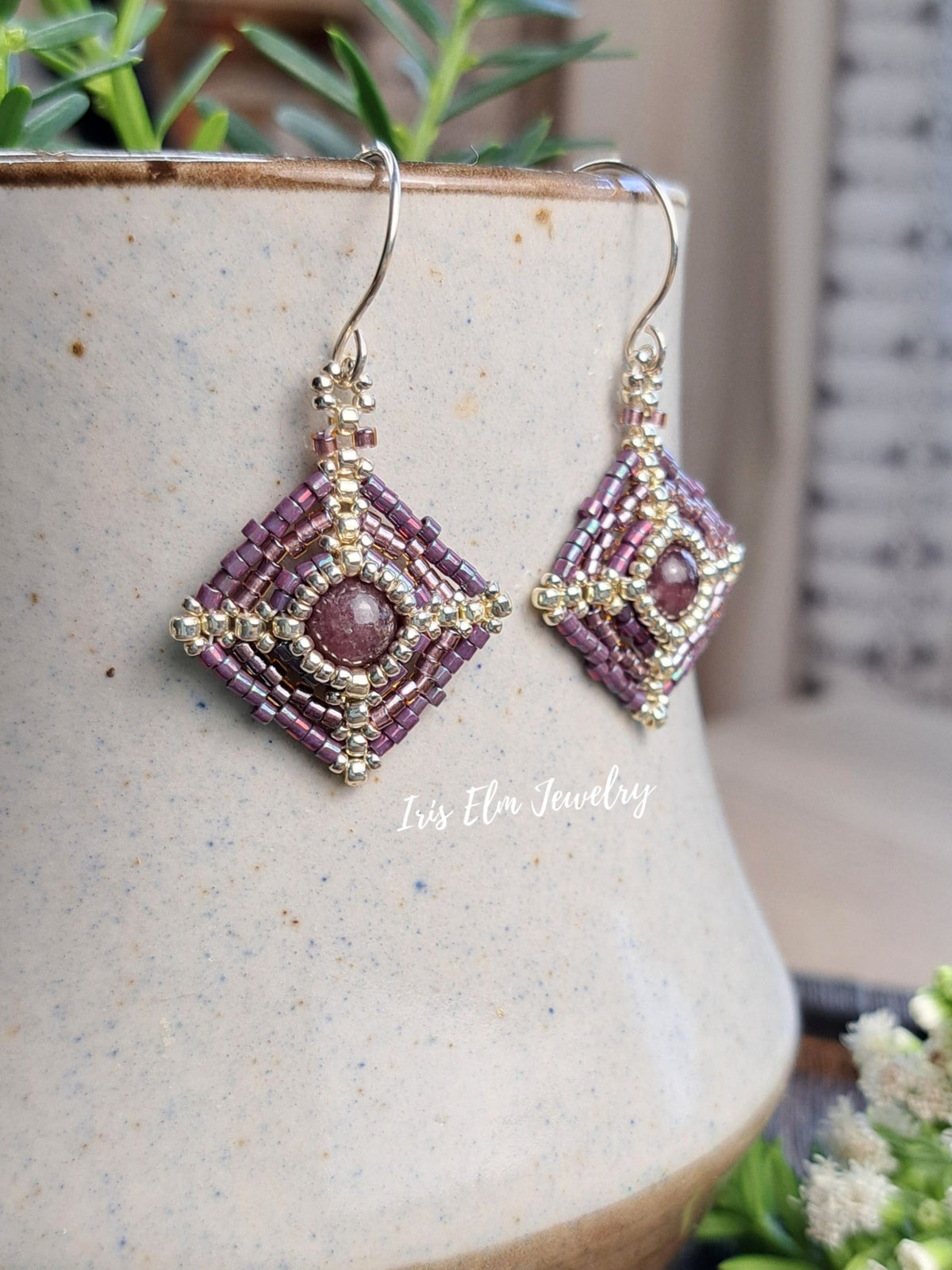 A pair of handmade beaded dangle earrings with purple and gold seed beads, hanging from silver fishhook wires. The earrings are displayed on a speckled ceramic pot with green foliage in the background.