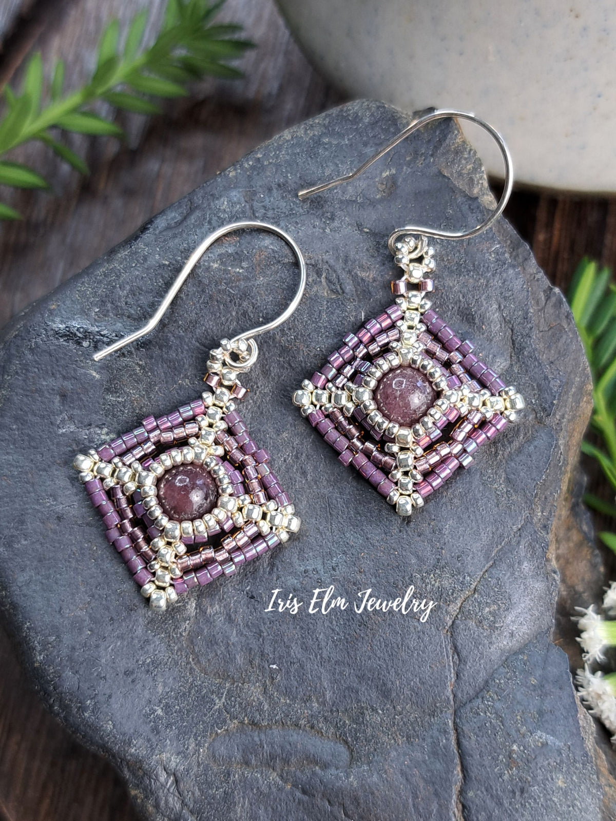 A pair of handmade square dangle earrings with silver-toned metal beadwork and purple seed beads, featuring a central oval purple stone. They are displayed on a dark gray rock with green foliage and a ceramic pot in the background.