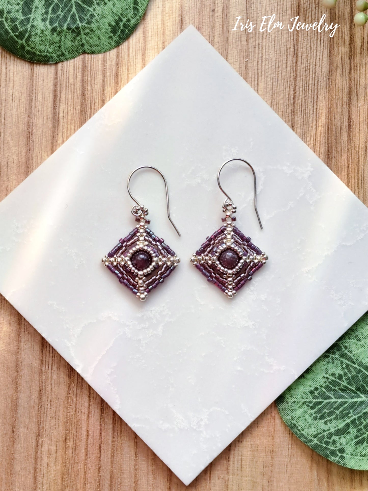 Pair of purple beaded lepidolite stone and seed bead earrings on a white tile with green leaves in the background, featuring 'Iris Elm Jewelry'.
