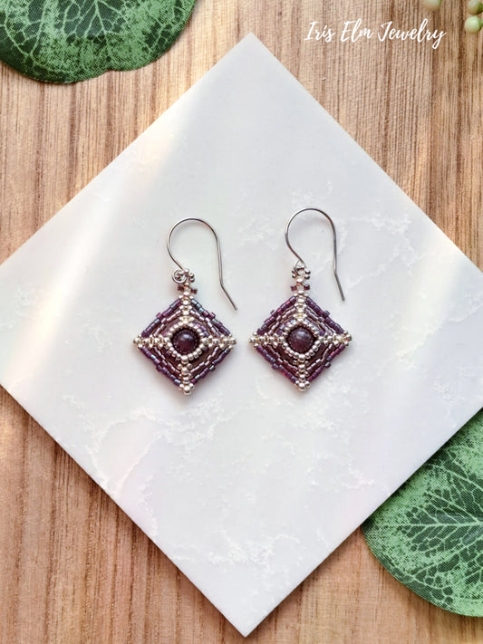Pair of purple beaded lepidolite stone and seed bead earrings on a white tile with green leaves in the background, featuring 'Iris Elm Jewelry'.