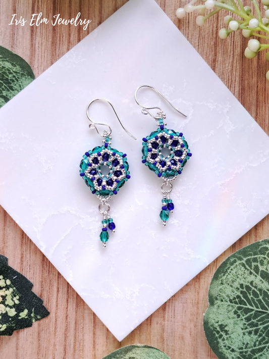 Pair of beaded cobalt blue and teal green earrings on a white card with 'Iris Elm Jewelry' branding, placed on a wooden surface with green leaves.