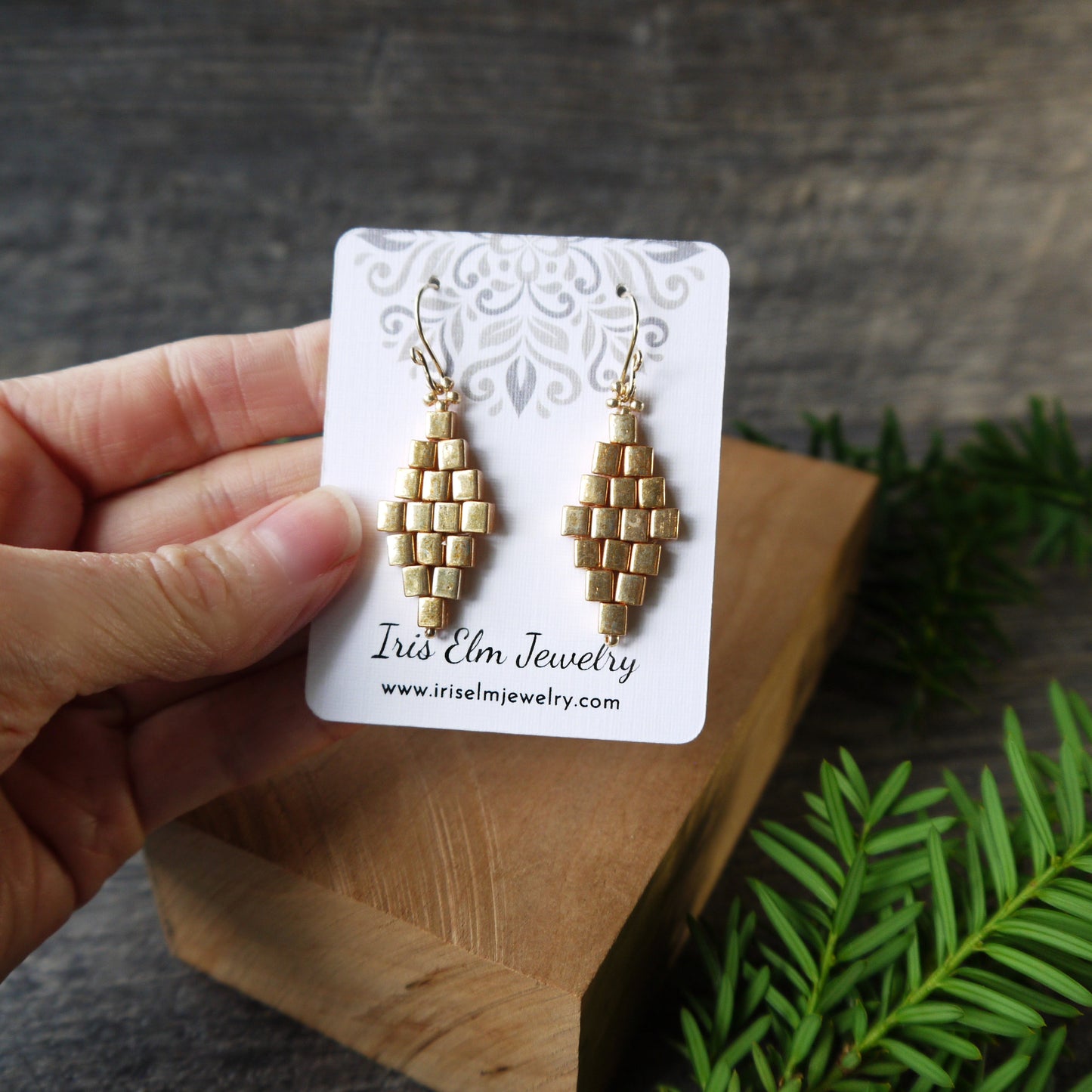 Gold beaded glass diamond earrings on a card held by a hand with a wooden surface and greenery in the background
