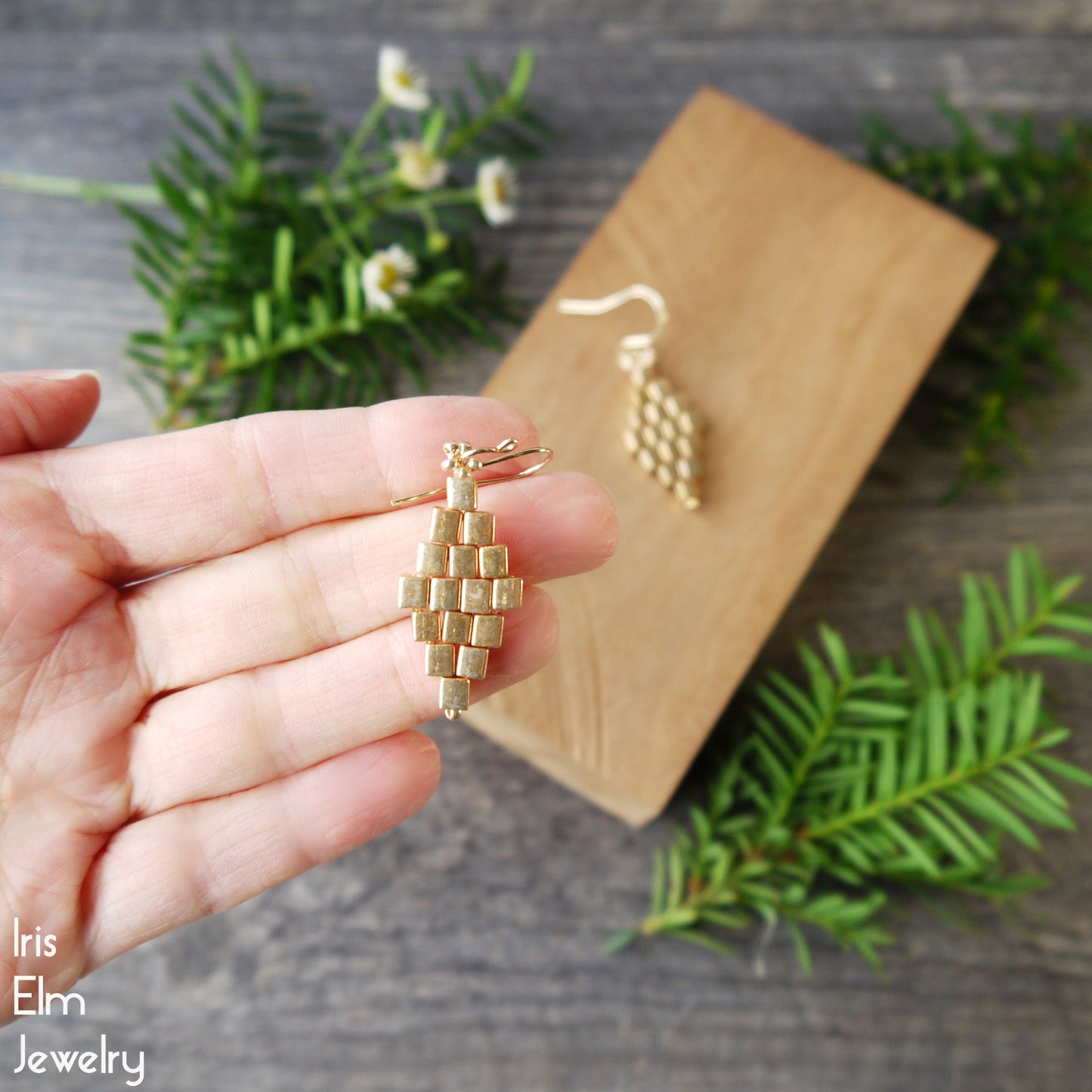 Gold beaded glass diamond shape earring held in hand with 'Iris Elm Jewelry' branding, wooden display block, and greenery in background.
