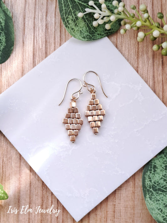 Pair of gold glass cube diamond woven earrings with 14k gold-filled ear wires on a white tile with green leaves in the background