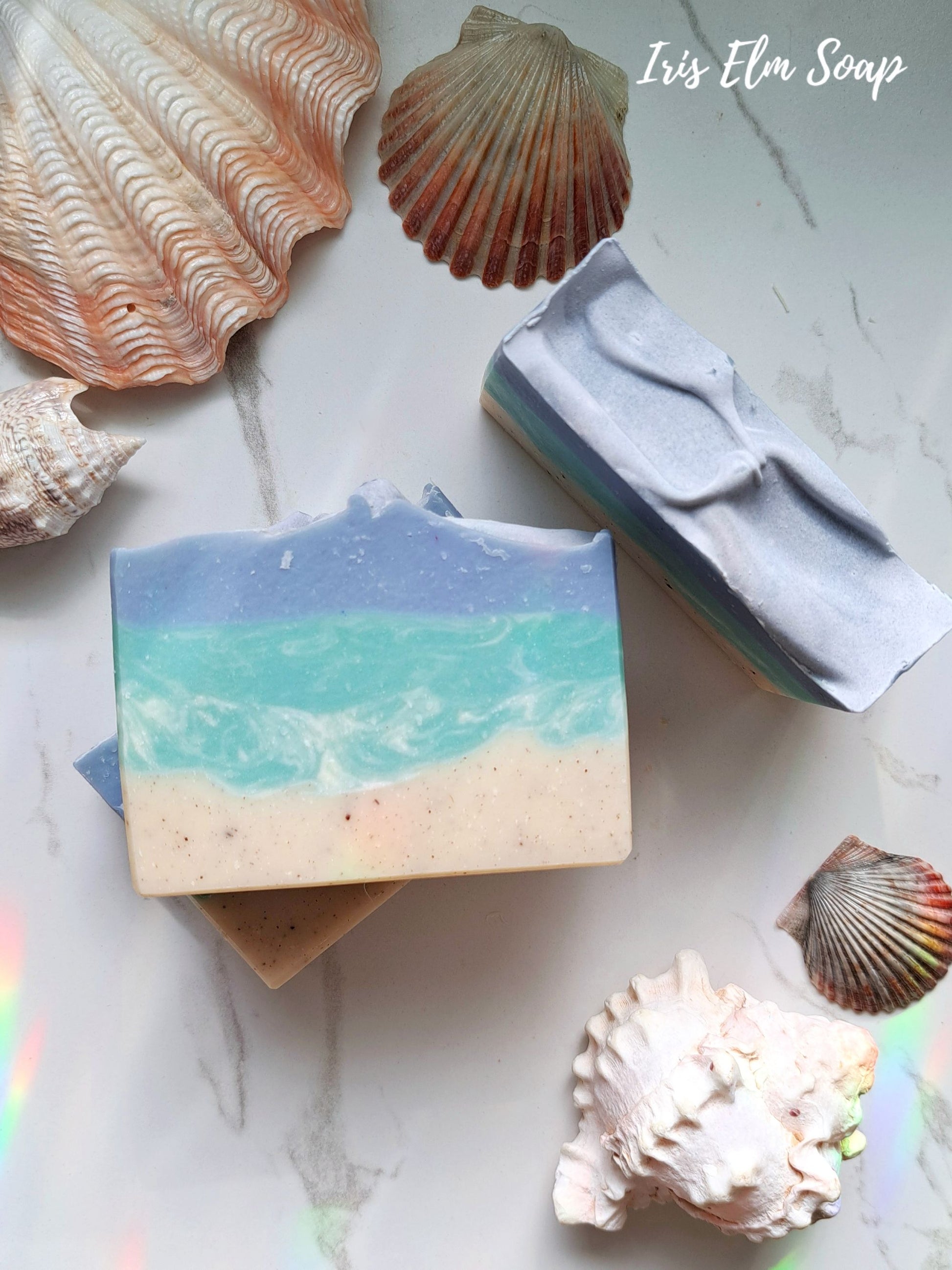 Two coconut lime verbena handmade soap bars with beach design surrounded by seashells on a light surface