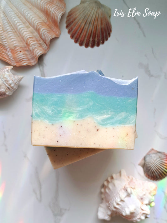 Bar of coconut lime verbena handmade soap with beach design surrounded by seashells on a marble surface