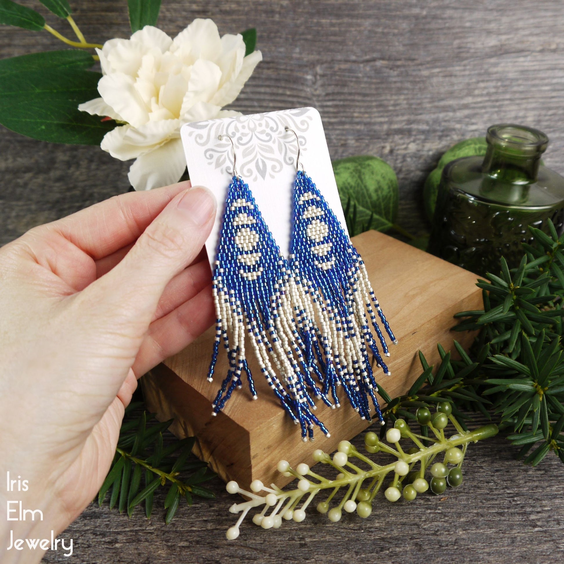 Blue and white beaded earrings held by a hand with floral and wooden background