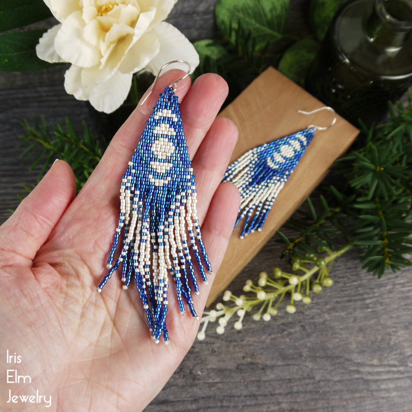 Beaded moon phase seed bead fringe handmade earrings held in a hand with a wooden box and floral background