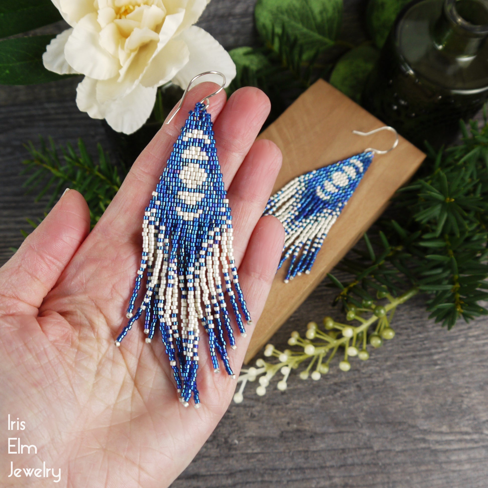 Beaded moon phase seed bead fringe handmade earrings held in a hand with a wooden box and floral background