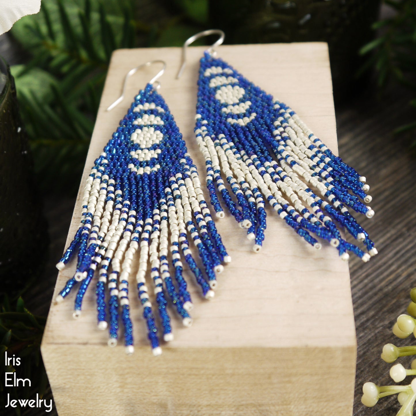 Blue and matte silver-white beaded moon phase fringe earrings on a wooden block with 'Iris Elm Jewelry' branding.