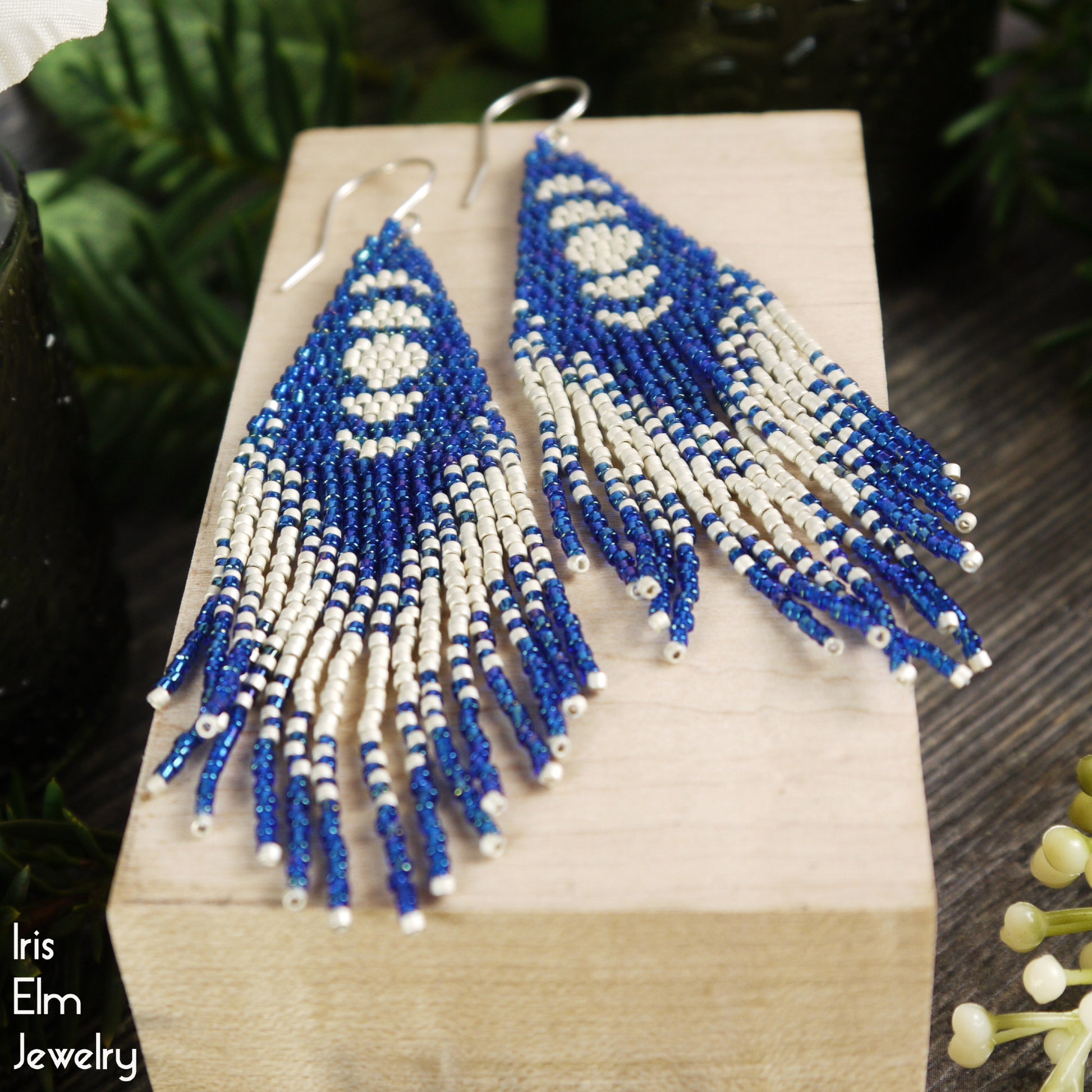 Blue and matte silver-white beaded moon phase fringe earrings on a wooden block with 'Iris Elm Jewelry' branding.