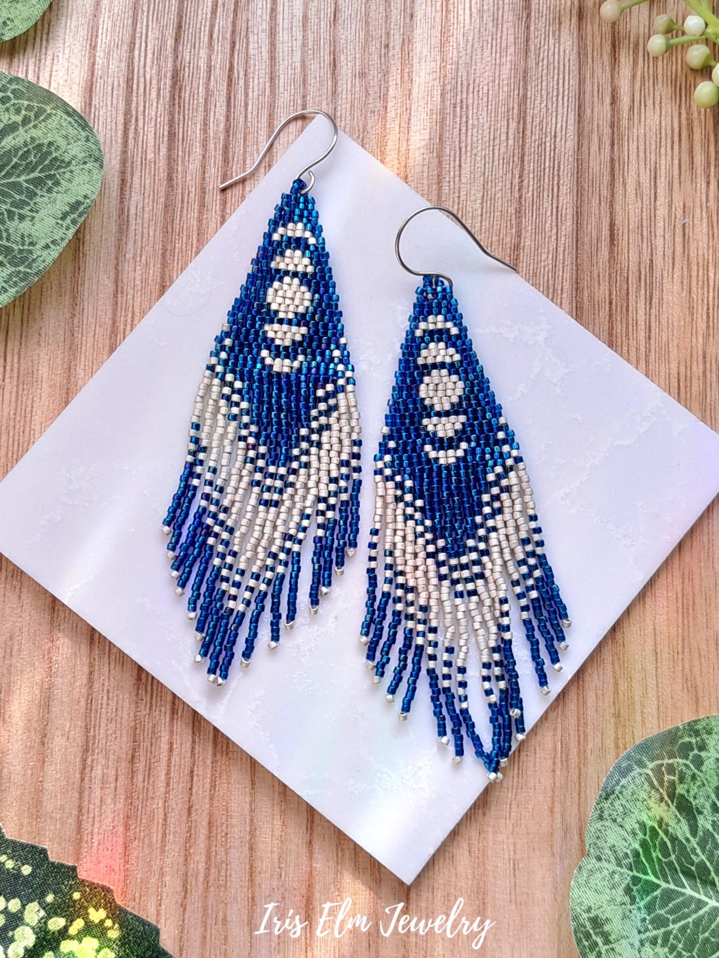 Blue and white beaded moon phase seed bead fringe earrings on a white card with a wooden background