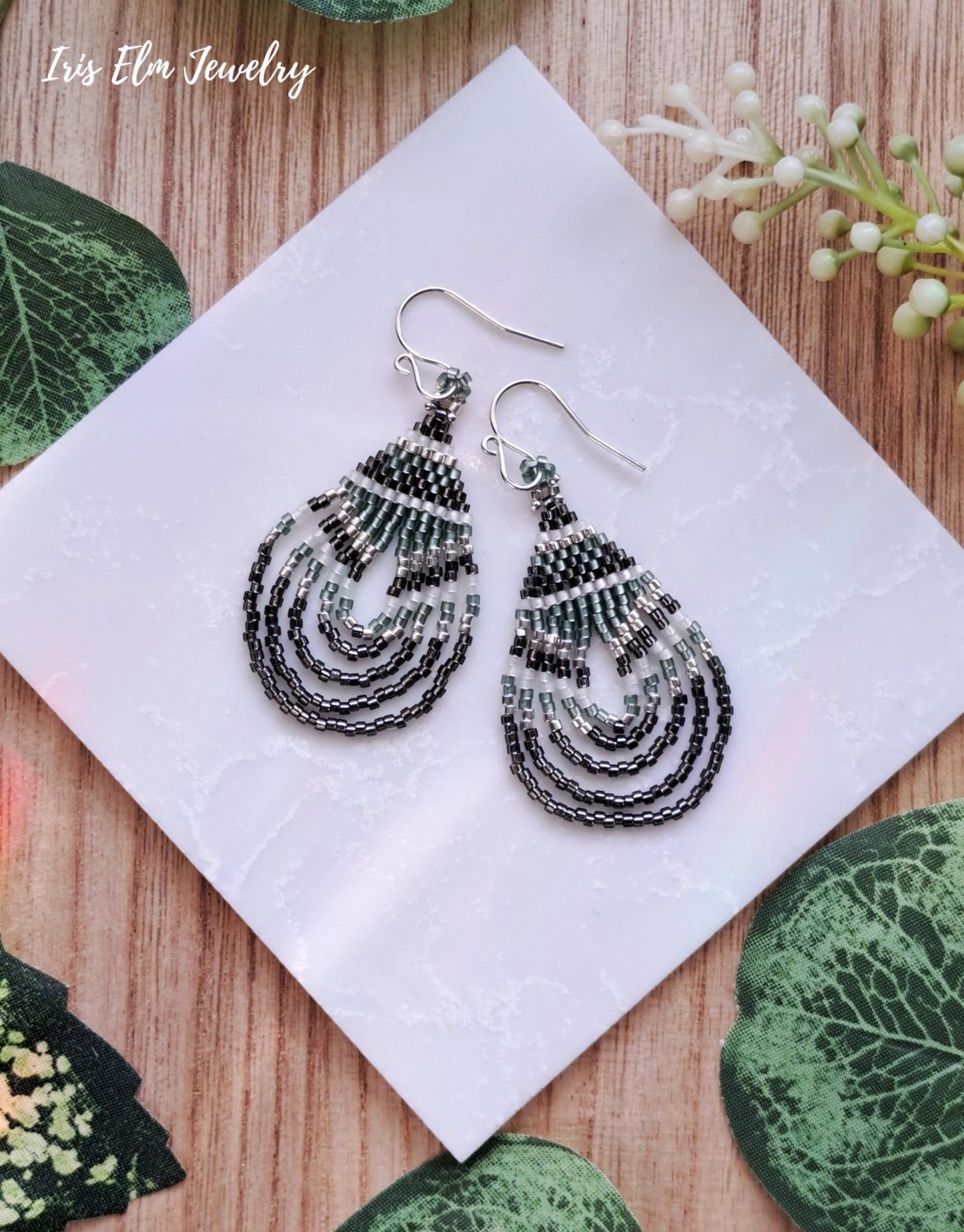Pair of beaded green and grey seed bead teardrop earrings on a white card with green leaves around