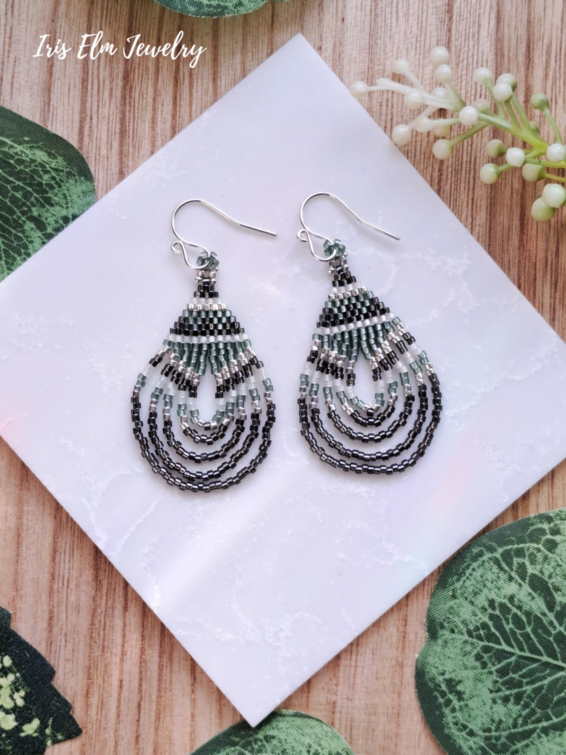 Pair of beaded green and grey seed bead boho teardrop earrings on a white card with 'Iris Elm Jewelry' branding, placed on a wooden surface with green leaves.