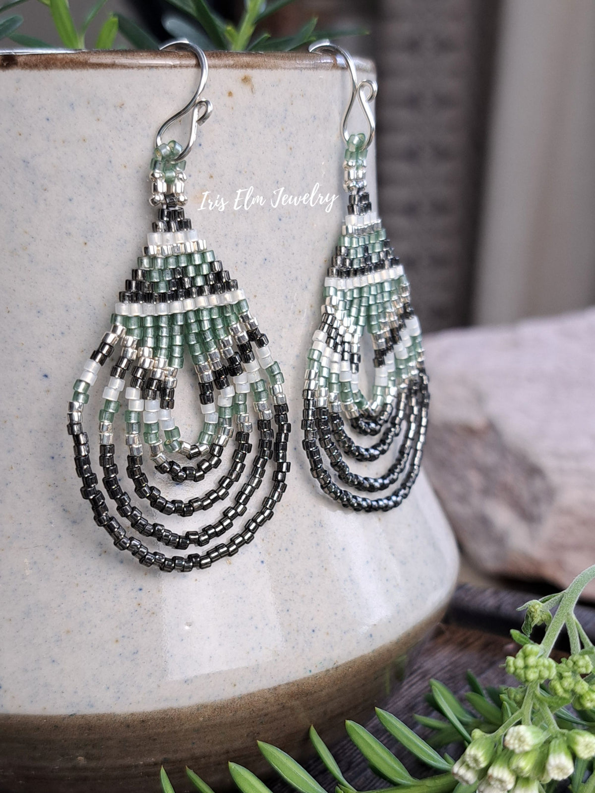A pair of handmade beaded dangle earrings with green, black, and white seed beads, hanging from silver fishhook wires on a ceramic vase. The watermark 'Iris Elm Jewelry' is visible on the upper left of the earrings.