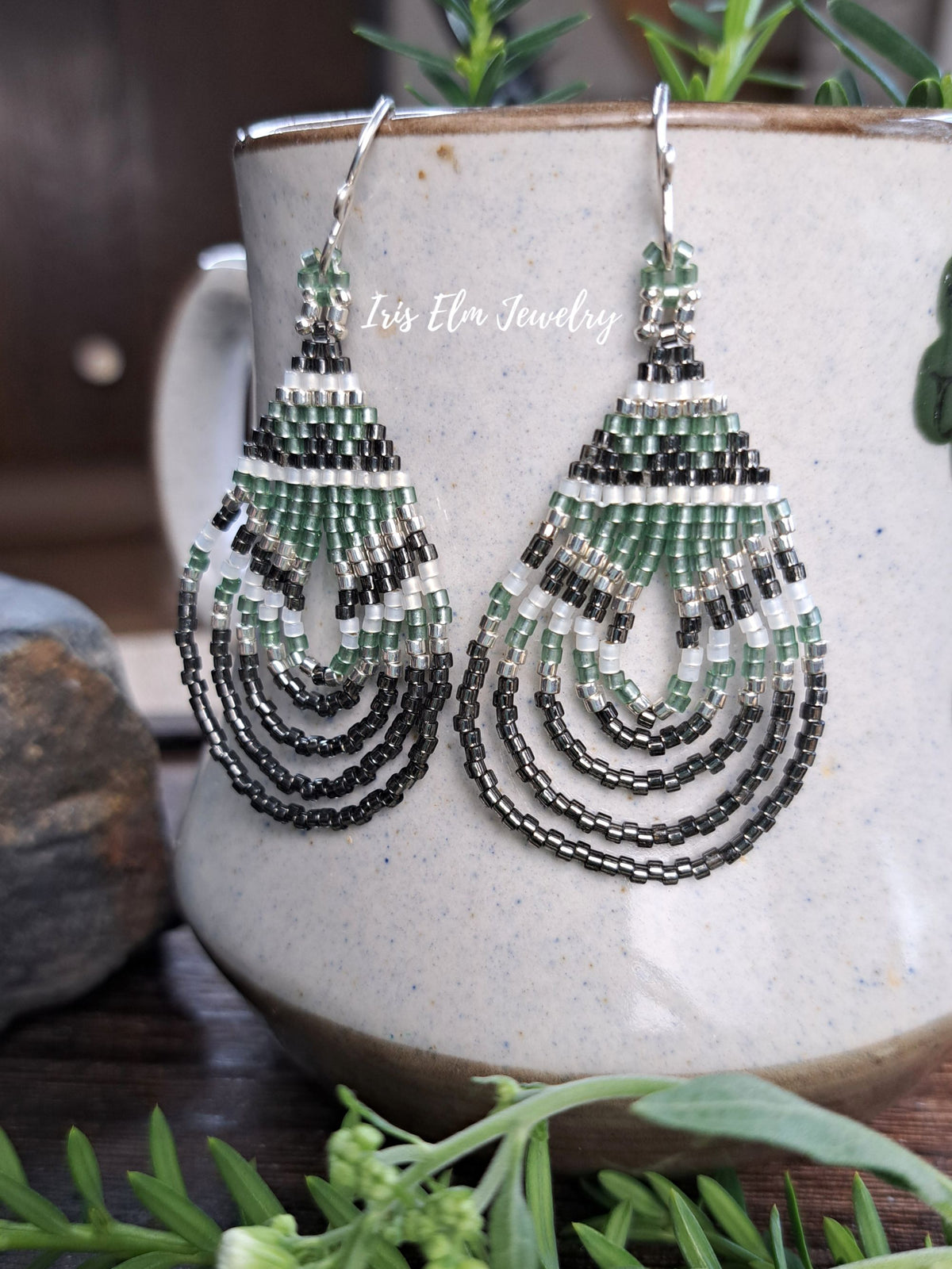 A pair of handmade beaded teardrop earrings with green, black, and white seed beads, hanging in front of a ceramic mug with a 'Iris Elm Jewelry' watermark.