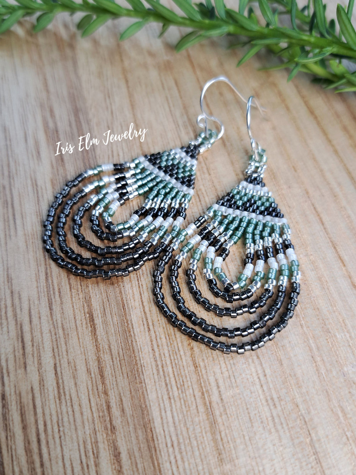 A pair of handmade beaded dangle earrings with silver fishhook wires, featuring concentric teardrop-shaped waves of black, green, and white seed beads. The earrings are displayed on a light wood grain surface with a sprig of green rosemary in the background.